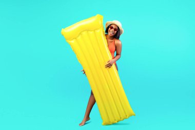 Şişirilebilir lilo tutan ve mavi stüdyo arka planında kameraya gülümseyen genç siyahi bir bayan. Havuzu yüzen Milenyum kadını tropik yaz tatilinde güneşli plaj gününün tadını çıkarıyor.