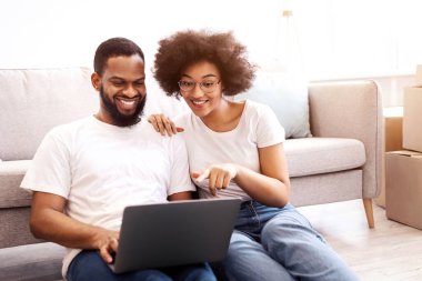 Siyah çift, taşındıktan sonra yeni ev için dizüstü bilgisayar alışverişi yapıyor, paketlenmiş taşınma kutularının arasında oturuyor. Yeni Daire Çevrimiçi Hizmetler, Gayrimenkul Satın Alma ve Sahiplik