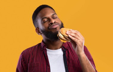 Afrikalı Milenyum Adamı Lezzetli Burger Sarı Stüdyo Arkaplanda Duruyor. Siyah adam hamburger yemekten zevk alıyor. Sağlıksız beslenme alışkanlığı, aşırı yeme. Abur cubur aşığı kavramı