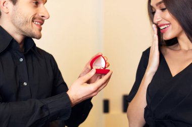 Will You Marry Me. Handsome bearded man holding and showing open box, offering marriage ring to his happy fiance, getting engaged in restaurant or at home during romantic date, closeup. Be My Wife