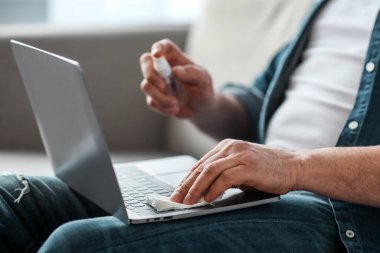 Erkek eller, Coronavirus salgını sırasında evde çalışırken dizüstü bilgisayarın klavyesini temizlemek için dezenfektan spreyi ve bez kullanıyor. Tanınmayan bir adam aletleri dezenfekte ediyor.