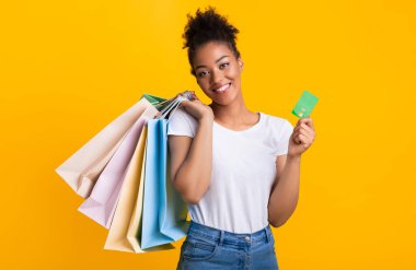 Alışveriş ve Nakit. Gülümseyen Afro-Amerikalı Kadın 'ın portresi kredi kartı ve parlak renkli alışveriş torbaları sarı arka plan duvarının üzerinde izole bir şekilde duruyor.