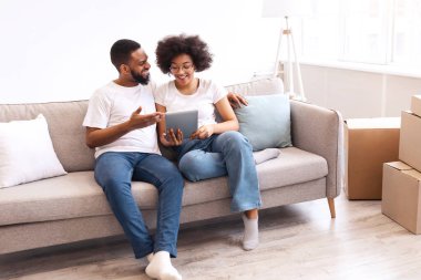 Yeni dairede taşındıktan sonra taşınan koltuklarda oturan mutlu siyah eşler tabletini kullanıyor. İnternette arama yapan çiftler taşınma işlemine hazırlanıyor. Boşluğu Kopyala