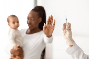 Çocuk Aşılarını Durdurun. Doktor şırıngayı tutuyor, anne hastayı reddediyor ve el işaretini avuç içi ile gösteriyor, elinde bebek var. Coronavirus aşısı, iğneye odaklan.