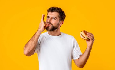 Lezzetli Fast food. Gülümseyen, Burger yiyen, parmaklarını yalayan, Sarı Stüdyo Duvarı 'nın üzerinde duran aç bir adamın portresi. Mutlu Gündelik Adam Atıştırmalık Adam Gülümseyerek Kameraya Bakıyor