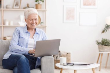 Yaşlılar için Online Derece. Mutlu yaşlı kadın evde dizüstü bilgisayarla çalışıyor, oturma odasında koltukta oturuyor, boşluğu kopyalıyor.