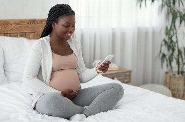 Mutlu Afrikalı genç hamile kadın evde akıllı telefonuyla rahatlıyor, sosyal ağlara bakıyor ya da hamilelik uygulamasını kullanıyor, yatak odasında oturuyor ve karnına dokunuyor,
