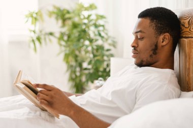 Siyah adam kitap okuyor. Evdeki yatak odasında rahat bir şekilde yatıyor. Afro-Amerikalı Adam, Hafta sonu Sabahları Rahatlayan En Çok Satanlar Kitabı 'nın tadını çıkarıyor. Daha Fazla Konsept Oku