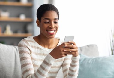 Mobil İletişim. Evde akıllı telefondan mesajlaşan genç siyahi kadın, oturma odasında dinlenirken arkadaşlarıyla cep telefonunda mesajlaşan neşeli Afrikalı Amerikalı kadın,