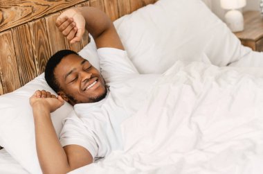 Mutlu Uykulu Siyah Adam, sabah uyandığında elleri uzanıyor, yatak odasında rahat bir şekilde uzanıyor, gözleri kapalı gülümsüyor. Günaydın Uyanış Konsepti. Yüksek Açı Görünümü