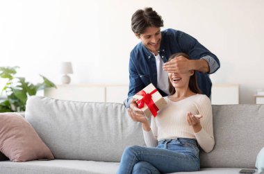 Gülümseyen genç Avrupalı koca karısına gözlerini kapar ve oturma odasının içinde hediye kutusu verir. Doğum günü sürprizi, sevgililer günü ve yıldönümü, Covid-19 karantinası sırasında evde romantik bir randevu.