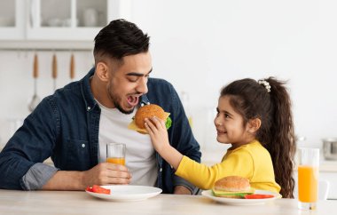 Küçük kızının, mutfakta birlikte yemek yerken, Arap babasını sandviçle beslemesi, eğlenmesi ve gülmesi, küçük tatlı bir kızın babasıyla eğlenmesi.,
