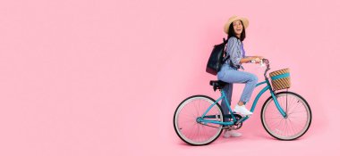 Yazlık şapka ve siyah sırt çantası takan heyecanlı Asyalı kadının uzun bir portresi hasır sepetli eski bir bisiklete biniyor ve pembe stüdyo arka planında izole edilmiş bedava fotokopi alanına bakıyor.