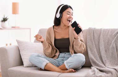 Mutlu Şarkıcı. En sevdiği şarkıyı söyleyen ve cep telefonunu kablosuz kulaklık takan Asyalı kadının portresi. Heyecanlı kadın evde eğleniyor, koltukta oturuyor, fotokopi çekiyor.