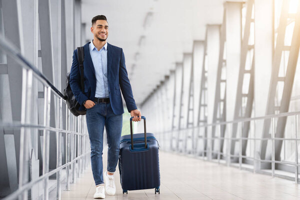 Готов к поездке. Красавчик араб, гуляющий с чемоданом в аэропорту, улыбающийся молодой человек с Ближнего Востока, несущий багаж во время полета к посадочным воротам, наслаждающийся предстоящим авиапутешествием