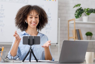 Covid-19 salgını sırasında öğretmenle İngilizce dersi videosu. Kablosuz kulaklıklı gülümseyen Afrikalı Amerikalı kadın oturma odasında dizüstü bilgisayarla web kamerasında video çekiyor.