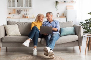 Cheerful Senior Couple Using Laptop Computer Sitting On Couch And Browsing Internet Together At Home. Mature People Use Gadget Watching Movie Online On Weekend. Full Length Shot