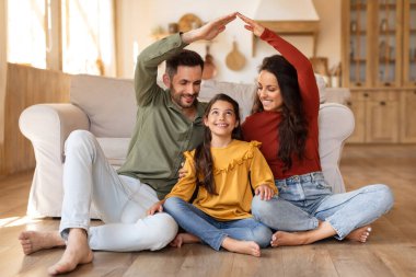 Gülümseyen aile, yerde otururken bir ev şekli oluşturarak iç açıcı bir sahne yaratıyor.