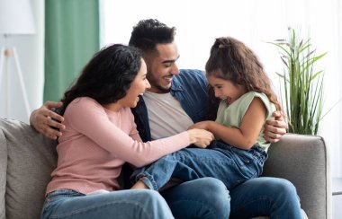 Üç kişilik neşeli bir Arap ailesi evde eğleniyor. Orta Doğulu ebeveynler küçük kızlarını gıdıklıyor ve oturma odasında dinlenirken birlikte gülüyorlar.