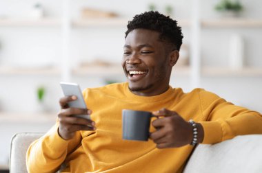 Mutlu Afro-Amerikan milenyumu çocuğu evdeki kanepede rahat rahat yaslanıyor, kahve içiyor ve modern akıllı telefon kullanıyor, kız arkadaşıyla sohbet ediyor, internette sörf yapıyor, uzayı kopyalıyor.