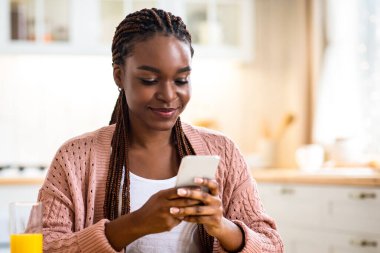 Gülümseyen Siyah Kadın, mutfakta kahvaltı ederken akıllı telefondan mesaj atıyor, neşeli Afrikalı Amerikalı kadın lezzetli yemeklerden zevk alıyor ve diyet uygulamasında kalorileri sayıyor, bedava uzay...