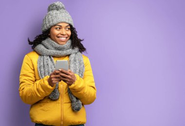 Mor Arkaplan Üzerine Akıllı Telefon Pozu Veren Kış Ceketi ve Şapkası Giyen Mutlu Siyah Kız. Stüdyo Çekimi, Boşluğu Kopyala