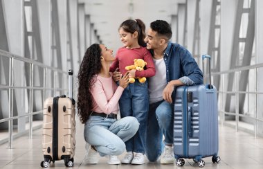 Havalimanında uçuşunu beklerken gülümseyen ve küçük kızlarıyla terminalde vakit geçiren üç kişilik mutlu bir Arap ailesi var.