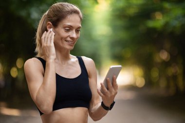 Pretty blonde middle aged athletic woman in black sportswear answering phone call while training outdoors at public park, pressing on earbud and looking at cell phone screen, copy space