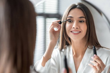 Makyaj Konsepti. Güzel genç bir kadın, banyoda hazırlanırken kirpiklerine maskara sürüyor, çekici bir kadın aynanın yanında duruyor, evde makyaj yapıyor, yakın çekim, seçici odaklanma.