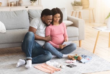 Neşeli hamile Afrikalı aile bebek kıyafetleri hazırlıyor ve liste yapıyor. Bebek doğurmaya hazırlanıyor. Oturma odasında yerde oturuyor.