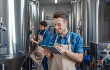 Bira fabrikasında kalite kontrol. Önlüklü iki gülümseyen adam çaydanlığın yanında tablet ve boş alan kullanarak çalışıyor.