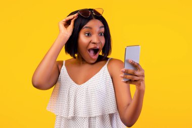 Yazlık kıyafetli siyahi kadın heyecanla akıllı telefona bakıyor turuncu stüdyo arka planında güneş gözlüğünü kaldırıyor. Şaşırmış Afrikalı Amerikalı kadın internette yaz indirimini öğreniyor.