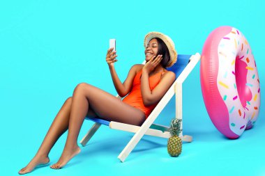 Çekici siyahi kadın şezlongda oturuyor, akıllı telefondan arkadaşıyla sohbet ediyor, mavi arka planda, afiş tasarımı. Yaz tatili boyunca internette iletişim kuran mutlu bayan, kopyalama alanı