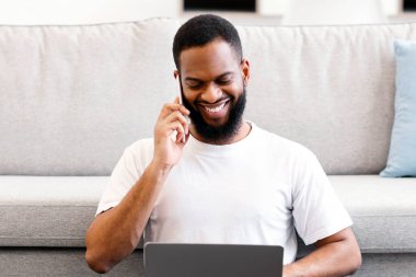 Telefonda konuşan siyahi serbest çalışan adam dizüstü bilgisayarını kullanıyor ve evde yerde oturuyor. Uzak Çalışma ve Uzak Mobil İletişim, Serbest Kariyer ve Modern Yaşam Tarzı