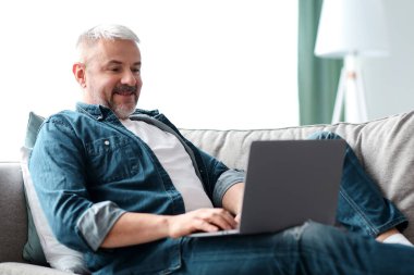 Oturma odasındaki koltukta dinlenirken dizüstü bilgisayar kullanan orta yaşlı, gri saçlı mutlu bir adam. Çevrimiçi blog okuyan, sohbet eden, internette sörf yapan neşeli, olgun bir adam.