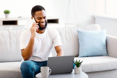 Cep telefonuyla konuşan Afrikalı adam evdeki koltukta oturan dizüstü bilgisayarla çalışıyor. Uzak Çalışma ve Modern Uzaktan İletişim, Serbest Yaşam Tarzı Konsepti