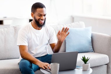 Siyah adam laptopta uzak bir aileyle video görüşmesi yapıyor, evdeki koltukta oturan bilgisayar kamerasına el sallıyor. Uzak Çevrimiçi İletişim Konsepti