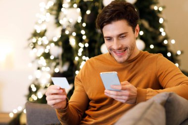 Smartphone kullanan gülümseyen milenyum adamı portresi, internet mağazasında internet üzerinden kredi kartı ödemesi, evdeki koltukta oturması. Kolay Dijital Para Aktarımı Konsepti