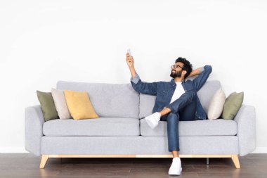 Rahat, sakallı, orta doğu sakallı ve gözlüklü genç adam evdeki koltukta oturuyor, uzaktan kumandayı tutuyor, AC 'yi açıyor, beyaz duvardaki kopyalama alanını işaret ediyor.