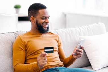 Afrikalı Amerikalı Adam kredi kartı ve akıllı telefon kullanarak işlem yapıyor ya da cep telefonu ile ödeme yapıyor. Evdeki koltukta oturuyor. Alışveriş Uygulaması ve E-Ticaret. Seçici Odaklanma