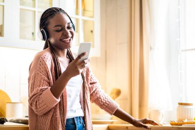 Akıcı Uygulama. Neşeli, genç Afrikalı, kablosuz kulaklık takan, akıllı telefonuyla müzik dinleyen, mutfaktaki iç kısımlarda dinlenen mutlu siyah bayan, favori şarkılarının keyfini çıkarıyor.