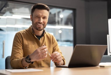 Modern Ofiste Çevrimiçi Çalışan Kameraya Gülümseyen Dizüstü Bilgisayarın yanında oturan Mutlu İşadamı. Girişimci Adam, işyerindeki masada oturuyor. İş ve Girişimcilik