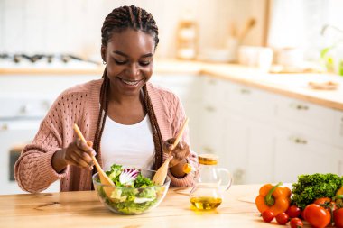 Sağlıklı Diyet Konsepti. Genç neşeli Afrikalı Amerikalı kadın mutfakta taze sebze salatası pişiriyor, gülen siyah kadın tabakta yeşillikleri karıştırıyor, evde öğle yemeğinin tadını çıkarıyor,