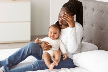 Doğum sonrası bunalım konsepti. Yorgun Afrikalı Amerikalı bir kadının portresi, küçük siyah çocuğuyla evde oturuyor, baş ağrısı çekiyor, alnına elle dokunuyor. Yorgunluk ve Ebeveynlik