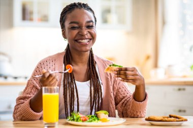 Güne Başlamak. Mutfakta lezzetli bir kahvaltı yapan mutlu siyah kadın, masada oturan neşeli Afrikalı genç Amerikalı kadın, sağlıklı sabah yemeğinin tadını çıkarıyor ve kameraya gülümsüyor,