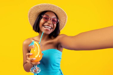 Havalı yaz kıyafetleri içinde güzel siyahi bir bayan ferahlatıcı bir kokteyl içiyor ve portakal rengi stüdyo arka planında selfie çekiyor. Genç ve mutlu bir kadın, tropikal tatilde kendi akıllı telefon fotoğrafını çekiyor.