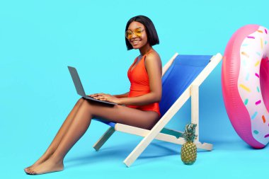 İnternette tropik cennetten çalışıyor. Bikinili mutlu siyah kadın şezlongda oturuyor ve mavi arka planda dizüstü bilgisayar kullanıyor. Afrikalı Amerikalı kadın internet üzerinden iş görüşmesi yapıyor, kopyalama alanı