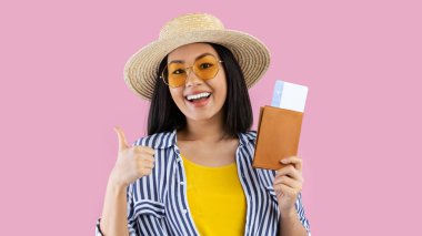 Tatil zamanı. Pasaportu ve biletleri olan heyecanlı Asyalı kadın başparmağını kaldırıyor, yaz gezisine hazır, hasır şapka ve gözlük takıyor, pembe stüdyo arka planında izole edilmiş, afiş takıyor.