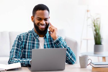 Genç siyahi iş adamı cep telefonuyla konuşuyor işyerinde oturuyor ve modern ofiste dizüstü bilgisayar kullanıyor. İş adamı telefonda biriyle sohbet ediyor. Mobil İletişim Konsepti