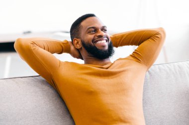 Mutlu Afro-Amerikalı adam evinde koltukta oturup ellerini başının arkasında tutarak rahatlıyor. Rahatlık ve rahatlık. Rahatlamış Adam Kanepede Dinleniyor Tembel Haftasonu Konseptinin tadını çıkarıyor